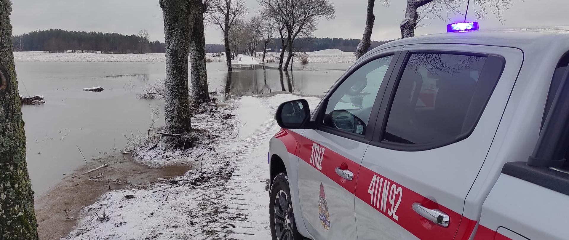 Zima. Z prawej strony na zdjęciu stoi samochód strażacki. dalej w oddali woda.