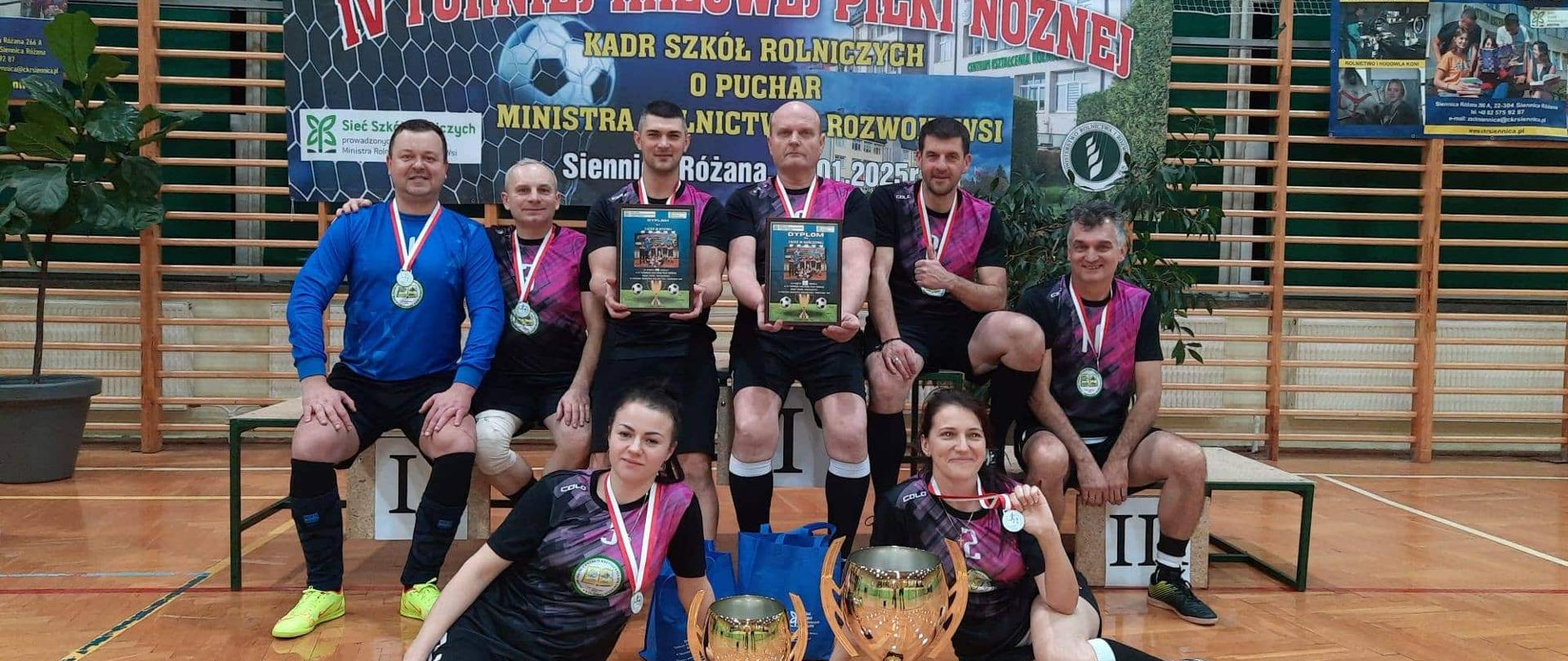 Drużyna w czarno-różowych strojach pozuje siedząc i stojąc na podium przed bannerem turniejowym; na pierwszym planie dwa złote puchary, zawodnicy trzymają dyplomy.