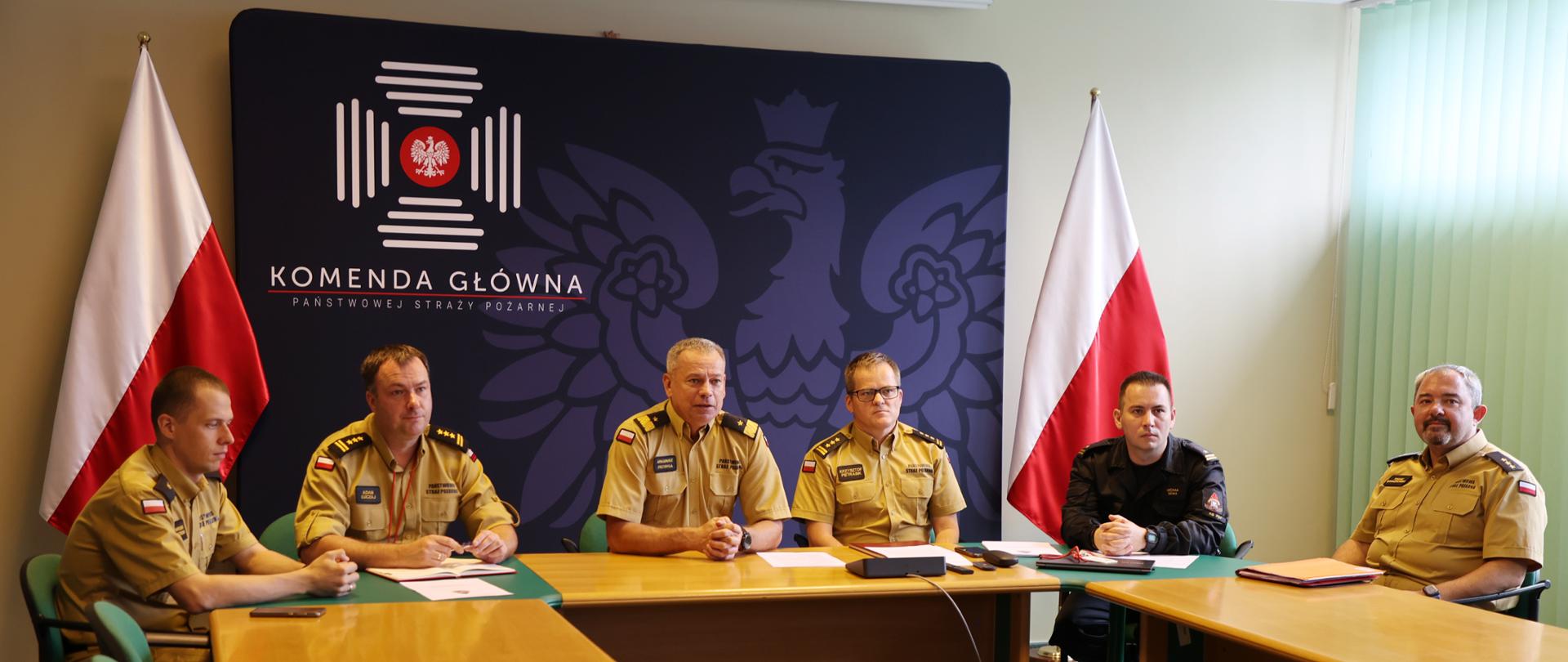 Pięciu mężczyzn w piaskowym umundurowaniu oraz jeden w czarnym siedzących przy stole. W tle baner Komendy Głównej Państwowej Straży Pożarnej oraz dwie biało-czerwone flagi