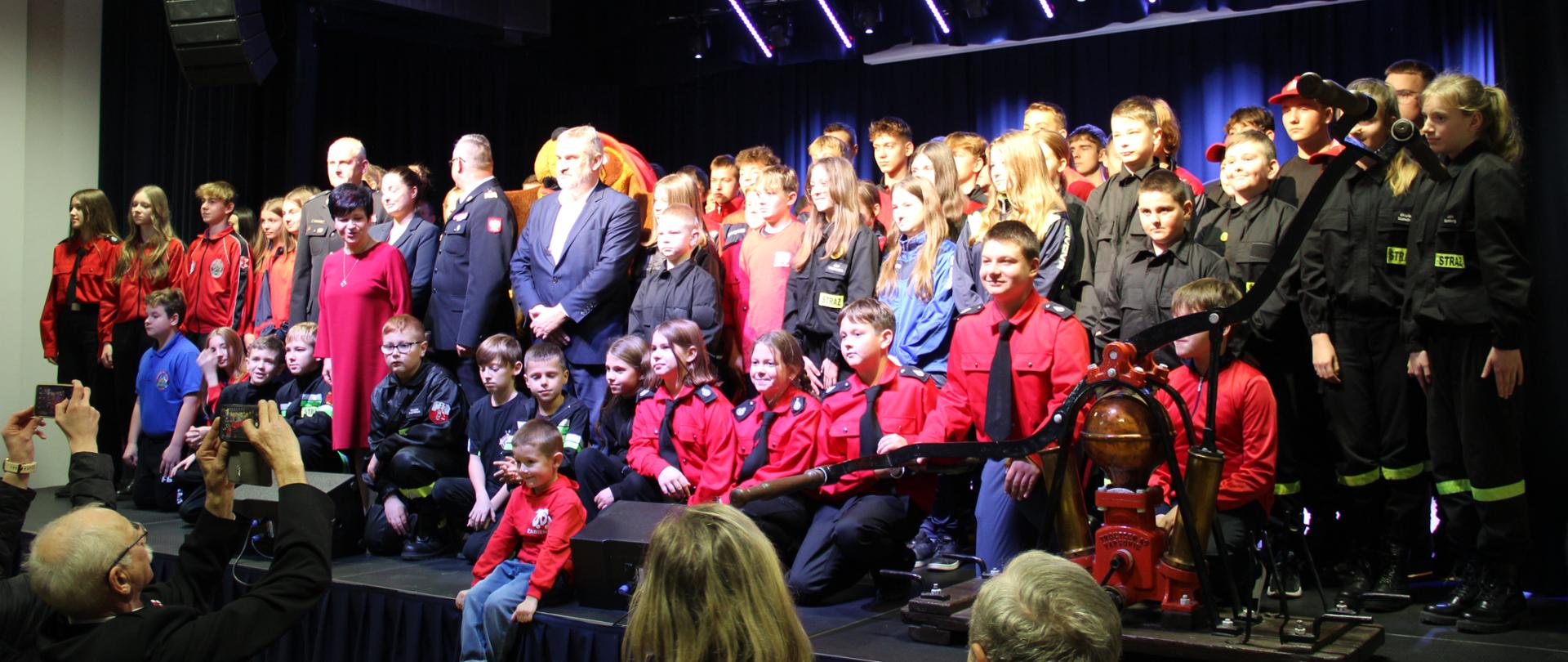 Zdjęcie grupowe młodzieżowych drużyn pożarniczych podczas odbierania promes. Kilkadziesiąt dziewcząt i chłopców stoją na scenie. Obojk nich stara pompa pożarnicza.
