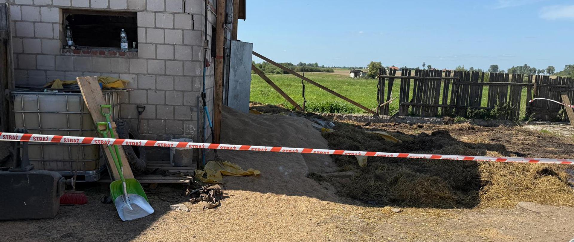 Róg budynku z pustaków, odgrodzony taśmą ostrzegawczą z napisem „STRAŻ”, w pobliżu narzędzia i sprzęty gospodarskie.