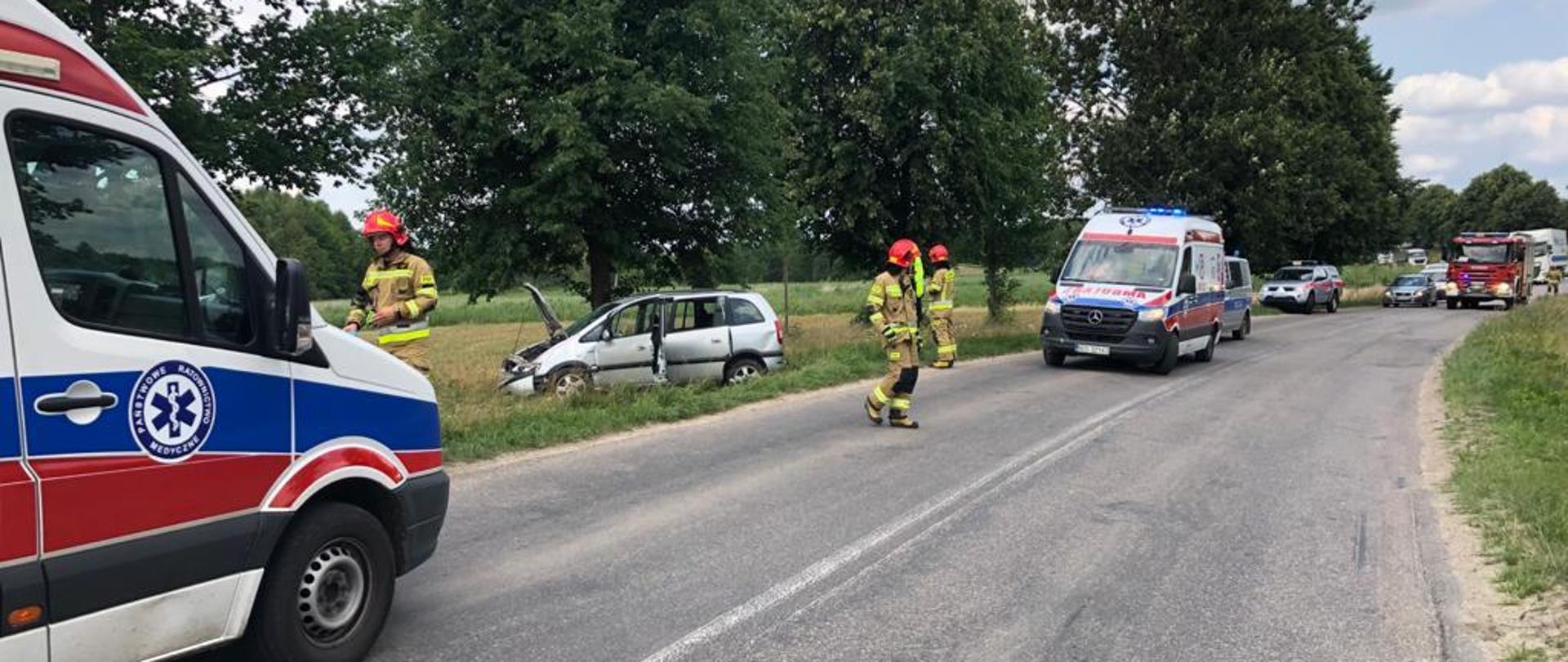 Rozbite auto osobowe stoi na poboczu. Strażacy przygotowują się do przyjęcia śmigłowca LPR