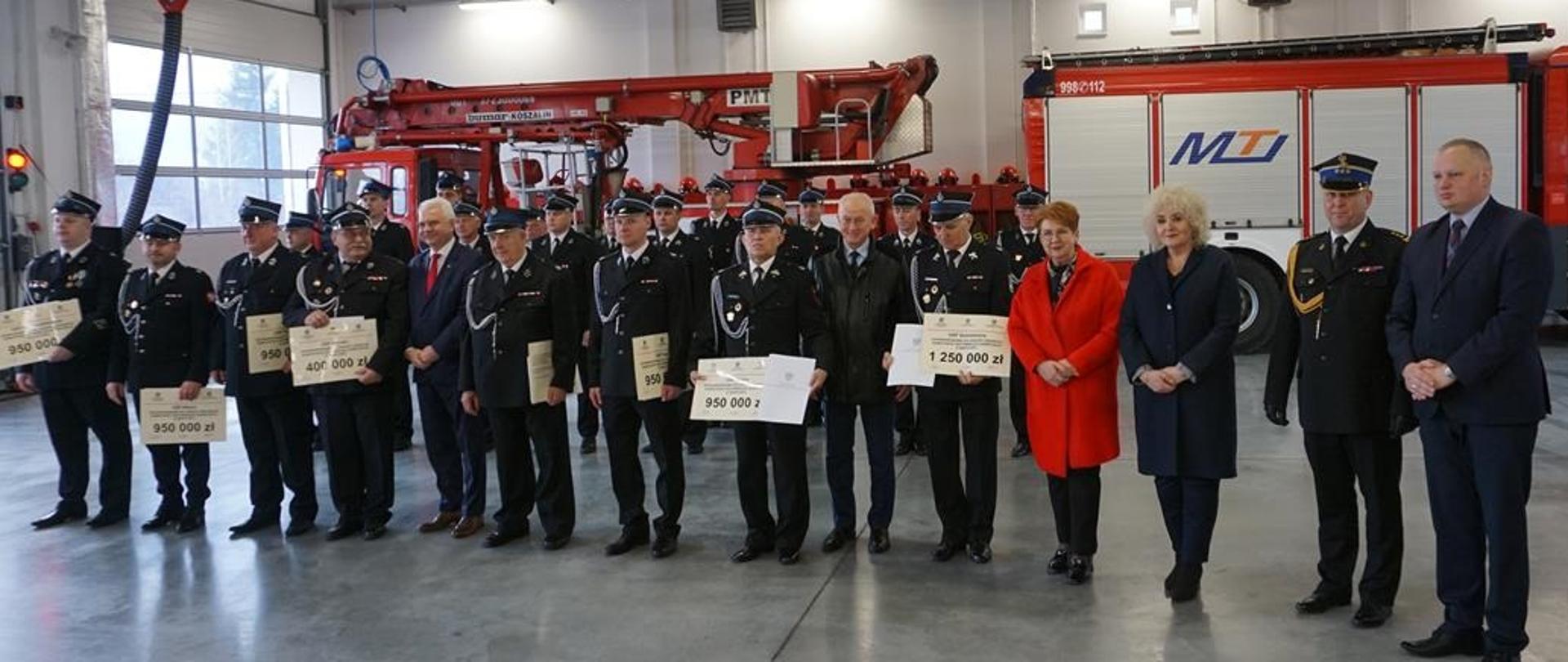 Prezesi OSP oraz zaproszeni goście stoją w szeregu, trzymają w rękach arkusze z kwotami dofinansowań.