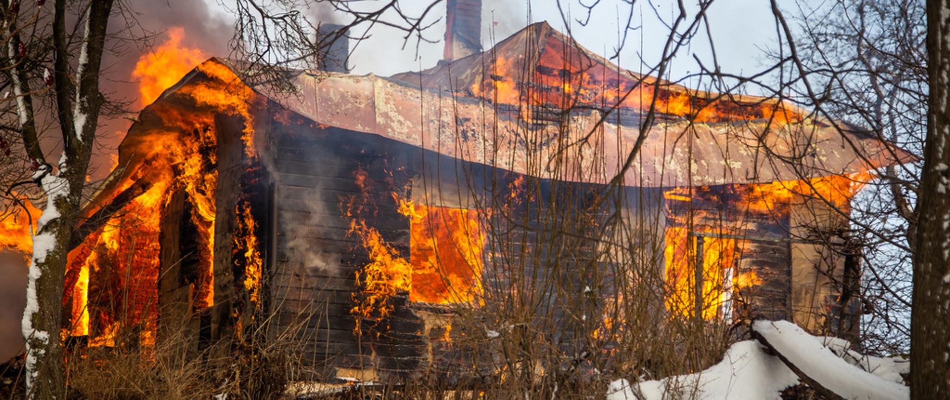 Pożar drewnianego budynku mieszkalnego. Budynek cały w ogniu z otworów okiennych drzwiowych i spod dachu wydobywa się ogień. Wokół domu teren zaśnieżony w pobliżu domu drzewa. 