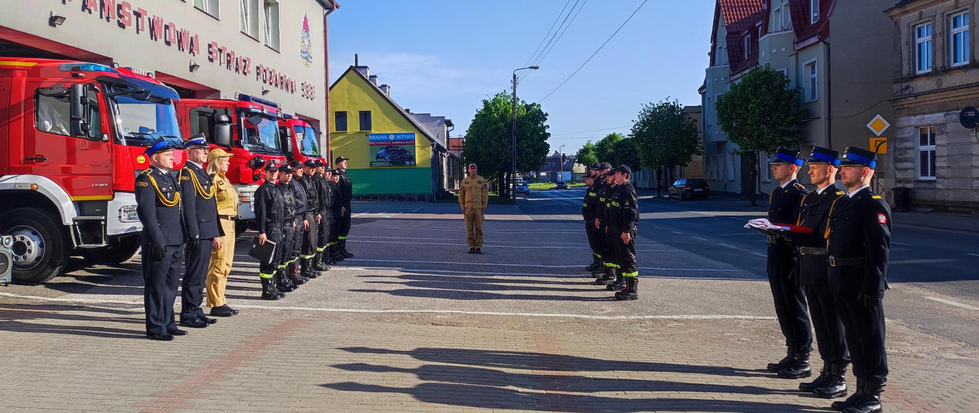 plac przed Komendą Powiatową Państwowej Straży Pożarnej w Sępólnie Krajeńskim, pracownicy Komendy, zmiany służbowe w ubraniach koszarowych wraz zastępcą Dowódcy Jednostki Ratowniczo-gaśniczej oraz poczet flagowy w mundurach galowych. Z lewej strony otwarte garaże i samochody ratowniczo gaśnicze.