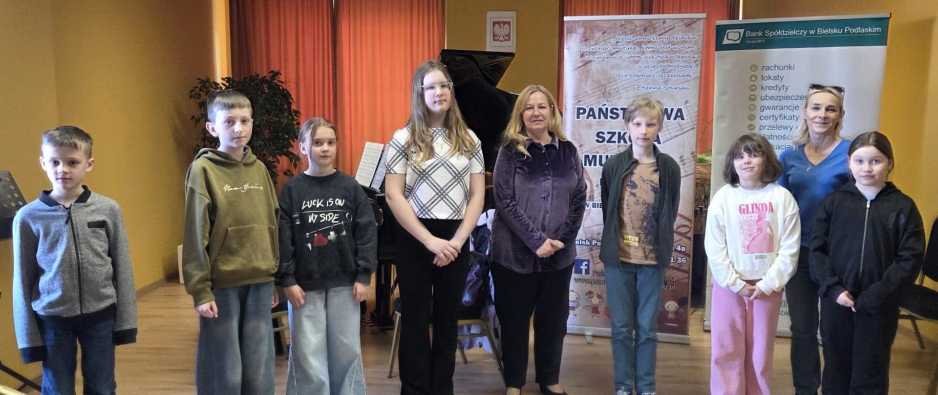 Na zdjęciu widzimy uczniów szkoły muzycznej klasy fortepianu na wspólnej fotografii w auli szkolnej. Po środku pani prowadząca warsztaty.