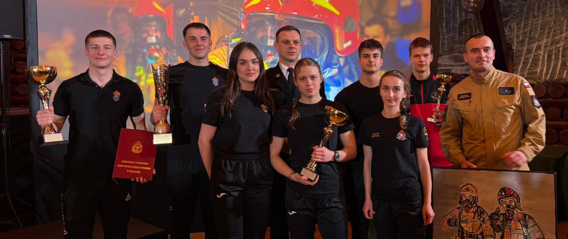 1. Grupa nagrodzonych drużyn
Zdjęcie grupowe przed podium, w dużej, oświetlonej komorze kopalni. Ośmioro młodych ludzi w jednolitych, czarnych strojach sportowych z dumą trzyma w rękach złote puchary i dyplom. Tło stanowi duży ekran z napisem "XIV Barbórkowy Turniej Strażaków i Ratowników...". Obok nich stoją dorośli mężczyźni: jeden w beżowym mundurze strażackim oraz jeden w mundurze galowym.

