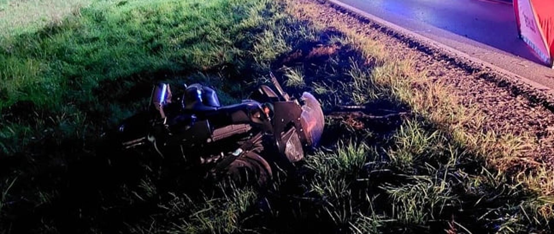 Na zdjęciu widoczny motor leżący na boku poza drogą i poboczem . W oddali widoczne samochody pożarnicze, karetka ZRM i strażacy. Wypadek w nocy , miejsce zdarzenia oświetlone przy użyciu najaśnic.