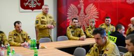 Na zdjęciu widać grupę strażaków z Państwowej Straży Pożarnej (PSP) w Polsce, którzy biorą udział w szkoleniu w sali konferencyjnej. Strażacy są ubrani w żółto-beżowe mundury z polskimi flagami na ramionach i różnymi oznaczeniami stopni oraz jednostek. Komendant Miejski PSP w Tarnowie stoi i przemawia do zgromadzonych, podczas gdy pozostali siedzą przy dużym drewnianym stole, słuchając go uważnie. Na stole znajdują się butelki z wodą mineralną, notatniki oraz kilka przekąsek.
Na ścianie w tle znajduje się duży herb Polski – biały orzeł w złotej koronie na czerwonym tle – oraz krzyż nad nim. Po prawej stronie widoczna jest czerwona ścianka z grafiką orła oraz napisem „PAŃSTWOWA STRAŻ POŻARNA KOMENDA MIEJSKA PAŃSTWOWEJ STRAŻY POŻARNEJ W TARNOWIE”, co wskazuje, że spotkanie odbywa się w jednostce PSP w Tarnowie.