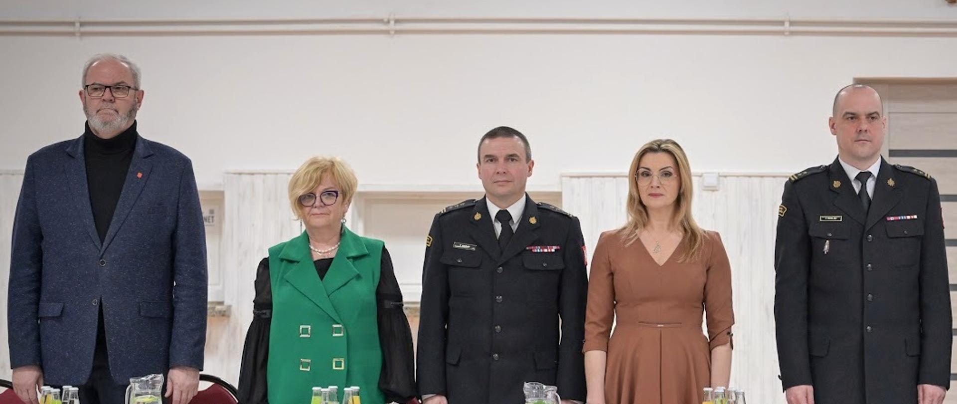 Zdjęcie zrobione w pomieszczeniu. Za stołem stoją uczestnicy spotkania, wśród nich dwóch oficerów Państwowej Straży Pożarnej w mundurach wyjściowych. Na stole ustawione są napoje oraz wizytówki z nazwiskami i funkcjami stojących osób.