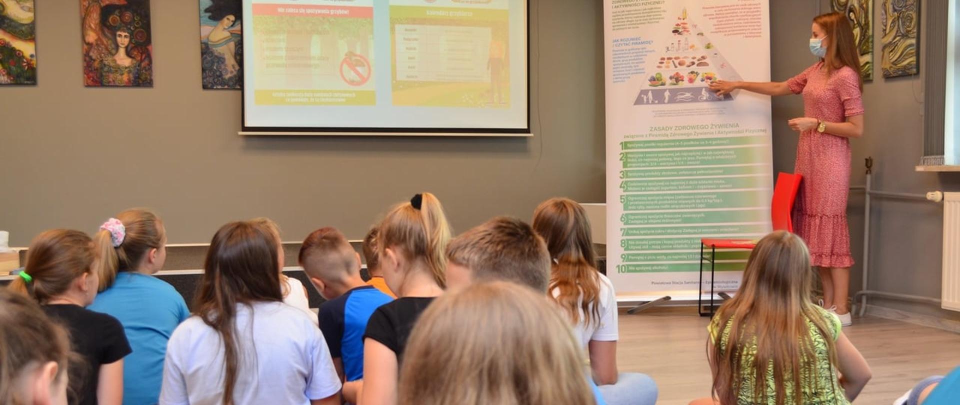Pani w maseczce stoi przy planszy przedstawiającej piramidę zdrowego żywienia i prowadzi wykład dla grupy dzieci 