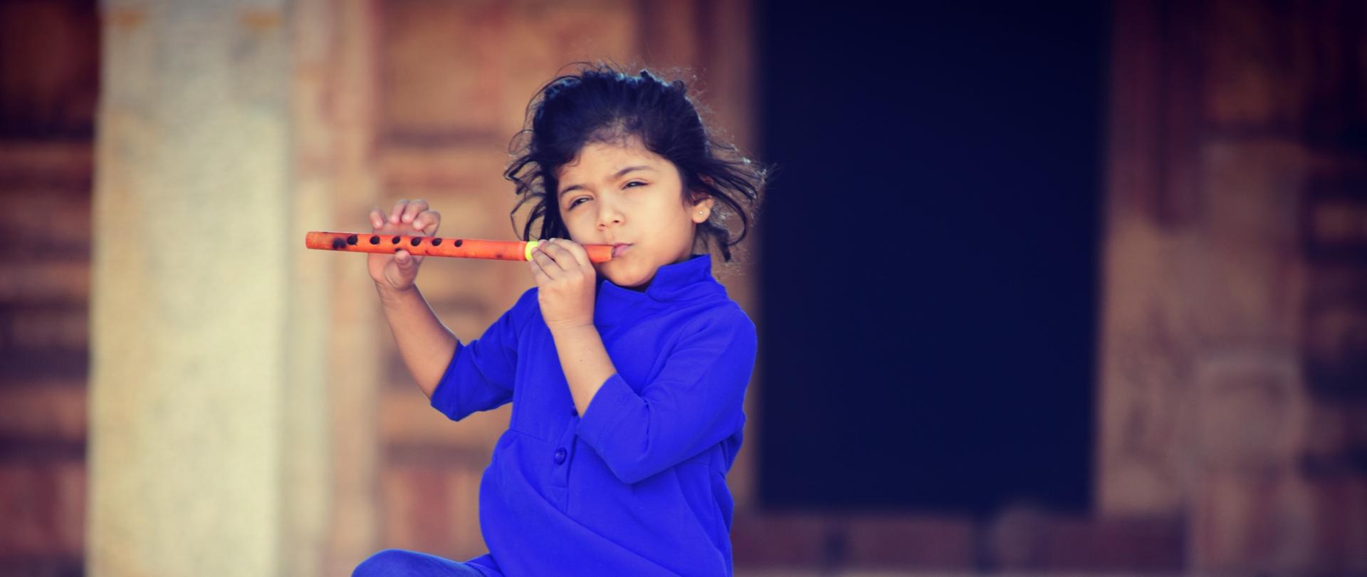 fotografia przedstawiająca ciemnowłose dziecko ubrane w niebieską tunikę i leginsy, siedzące na schodach i grające na flecie