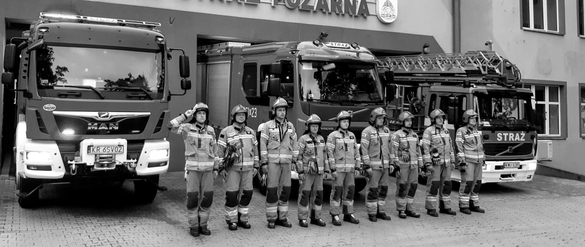 Na zdjęciu strażacy w ubraniu specjalnym stojący w szyku przed samochodami ratowniczo gaśniczymi przed budynkiem Komendy Powiatowej PSP w Wieliczce