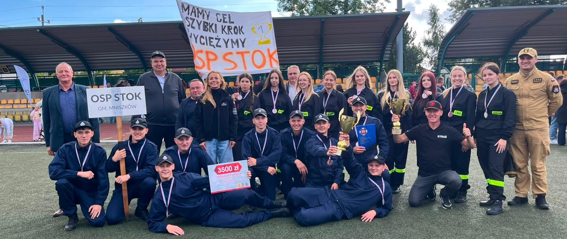 Grupowe zdjęcie uśmiechniętych drużyn strażackich OSP Radwan oraz OSP Stok, pozujących na boisku sportowym po VII wojewódzkich zawodach sportowo-pożarnicze Ochotniczych Straży Pożarnych z terenu województwa łódzkiego. Kobiety oraz mężczyźni, którzy brali udział w zawodach ubrani są w ciemne mundury strażackie, na szyjach mają zawieszone medale. Strażacy trzymają w rękach puchary za osiągnięte miejsce, dyplomy oraz czek z nagroda pieniężną. Z prawej strony grupy stoi Komendant Powiatowy PSP w Opocznie, natomiast z lewej strony prezesi drużyn oraz Pani Burmistrz gminy Białaczów. Cała grupa pozuje z uśmiechem w świetle dziennym n a tle infrastruktury sportowej oraz drzew.
