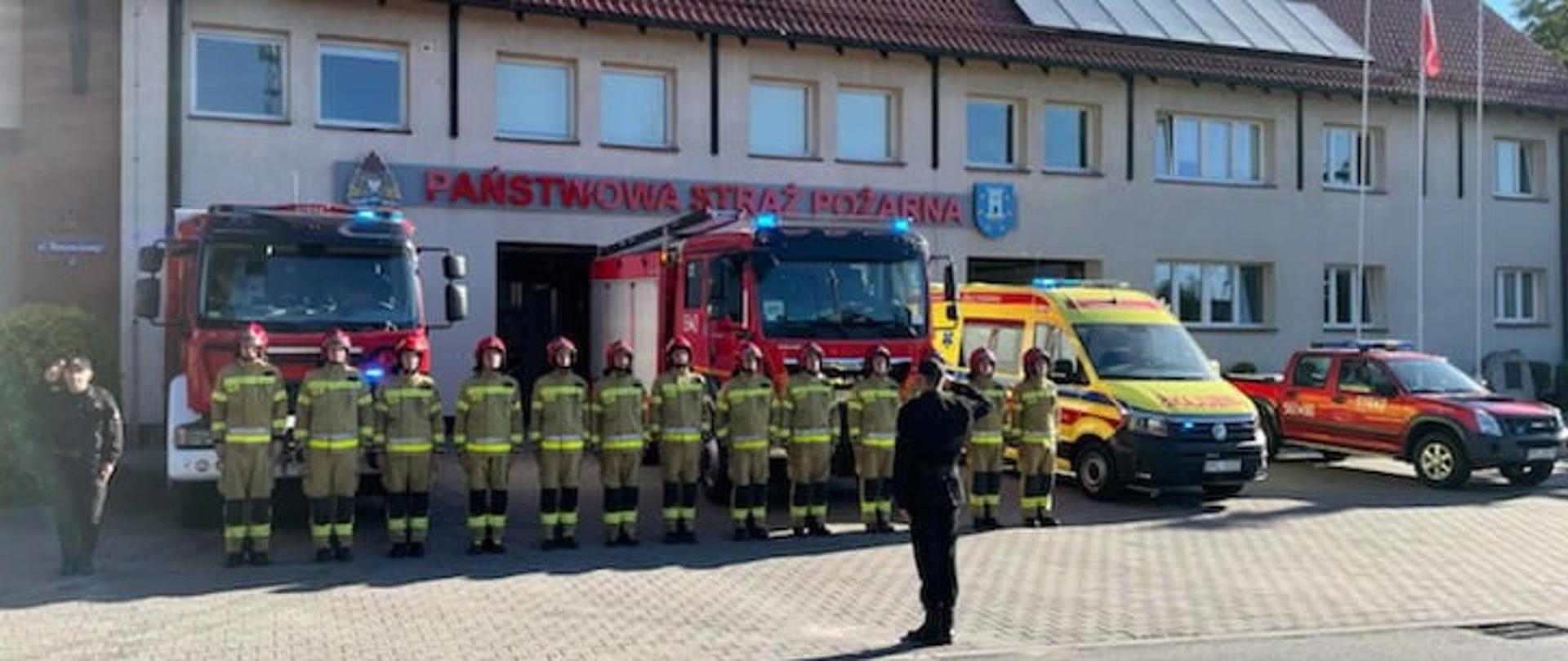 Front budynku straży pozarnej, przed nim stoją samochody gaśnicze oraz karetka medyczna, na pierwszym planie stojący w jednym szeregu strażacy ubrani w ubrania specjalne , z lewej strony szeregu i przed nim stoją salutujący strażacy ubrani w czarne mundury sztabowe PSP.