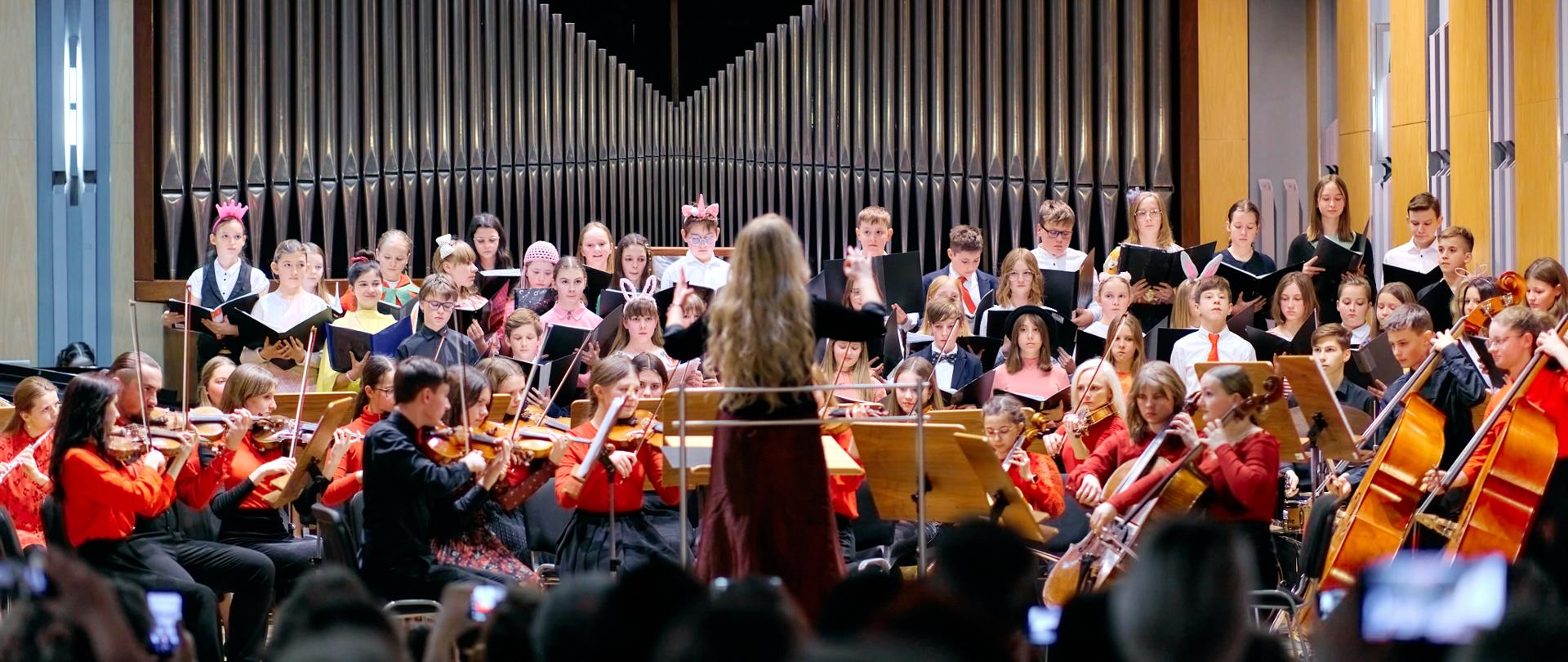 Na pierwszym planie publiczność. Na środku sceny orkiestra z dyrygentem. Orkiestra ubrana w kolorze czerwono- czarnym. Powyżej uczniowie chóru dziecięcego ubrani kolorowo trzymający w ręku nuty w czarnych teczkach. W tle organy