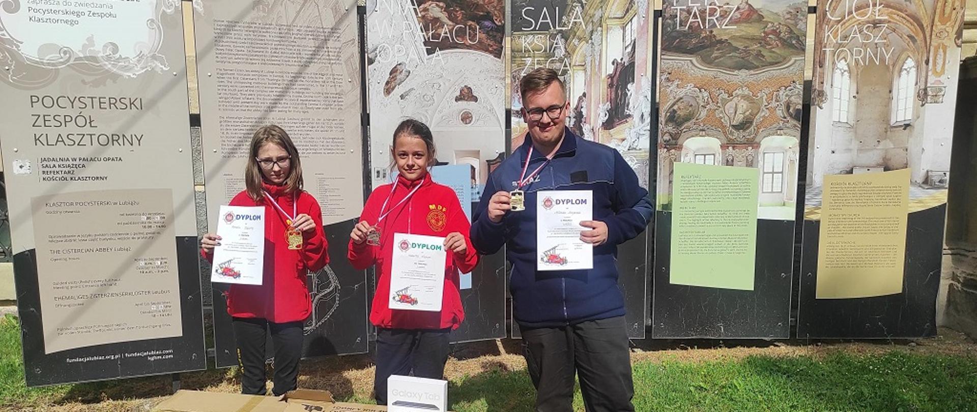 Dzień. Na tle tablic informacyjnych stoją trzy osoby - od lewej dwoje dzieci, z prawej młody mężczyzna. Od lewej dwójka ubrana w czerwone bluzy i czarne spodnie. Z prawej chłopiec ubrany w spodnie koszarowe w kolorze czarnym oraz żółte elementy odblaskowe na nogawkach oraz niebieską bluzę polarową. Dzieci trzymają w jednej ręce dyplom a w drugiej prezentują medale wiszące im na szyi. Przed nimi na trawie znajdują się dwa pudła kartonowe w kolorze szarym . Na pudłach widnieje rysunek złożonej hulajnogi elektrycznej.