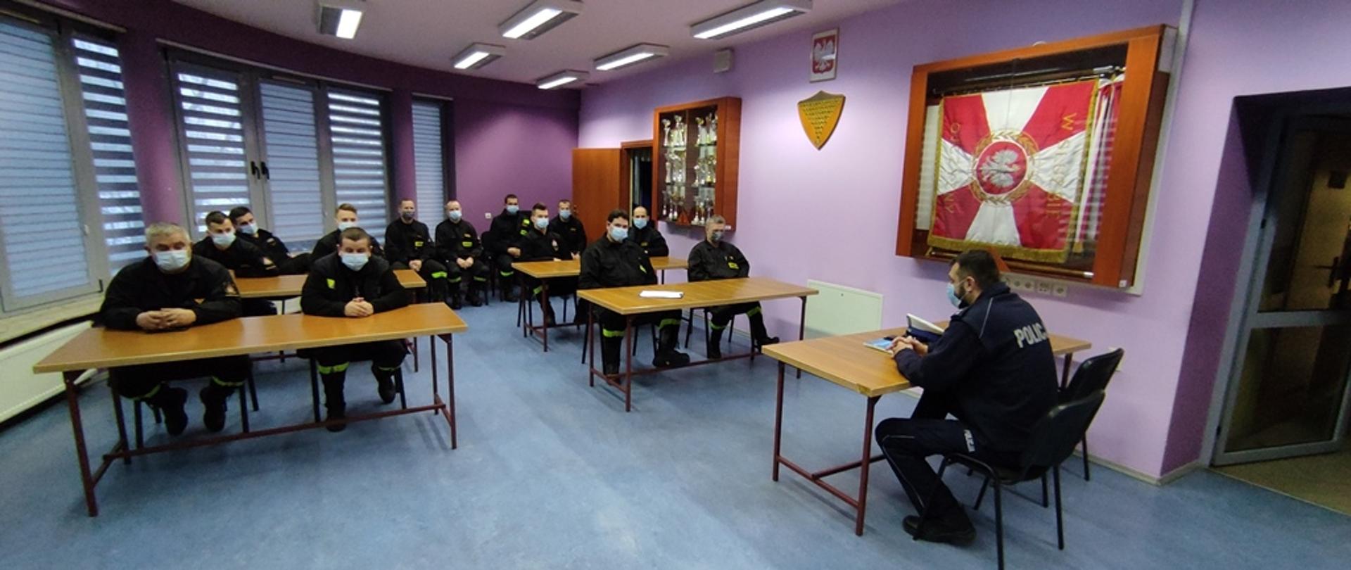 Sala Narad Komendy Powiatowej PSP we Włoszczowie, strażacy uczestniczący w szkoleniu w umundurowaniu koszarowym siedzą przy stolikach, frontem do nich siedzi policjant prowadzący szkolenie. 