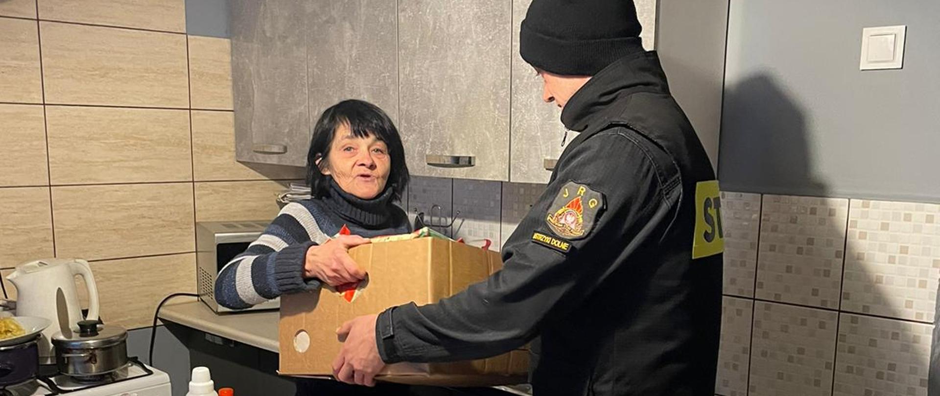 Na zdjęciu widać strażaka PSP w ubraniu koszarowym który przekazuje zakupione dary dla potrzebującej kobiety. Jedno pudło strażak przekazuje kobiecie w jej ręce. Na stole stoi więcej darów. 