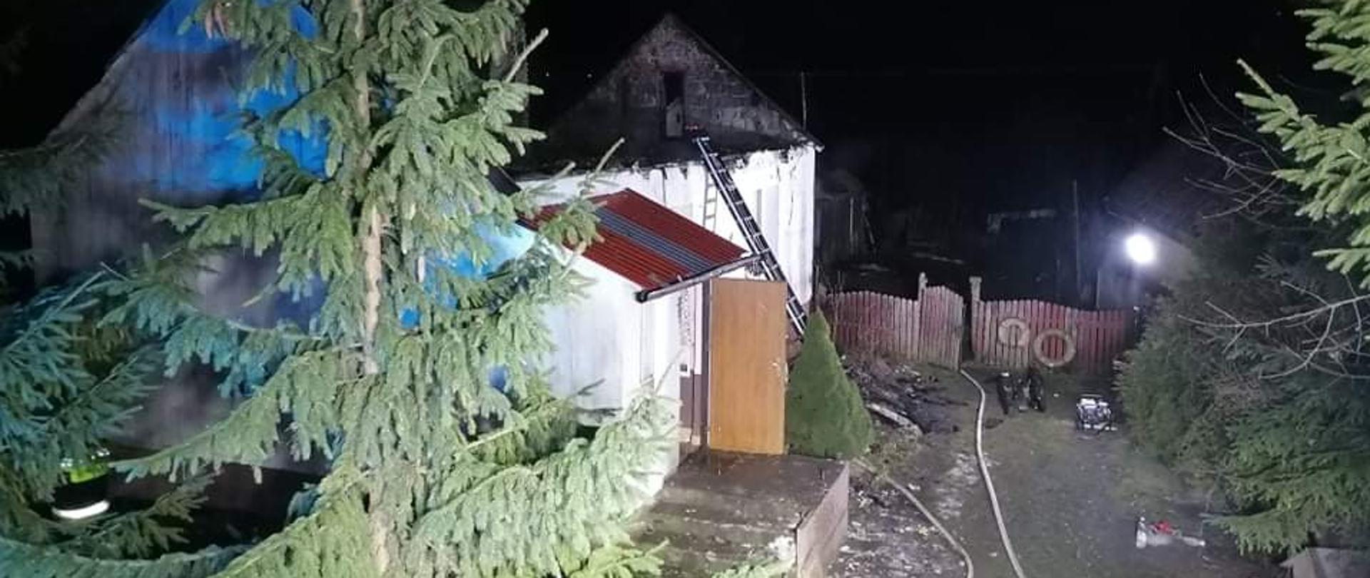 Zdjęcie przedstawia posesję gdzie w domu mieszkalnym wybuchł pożar. Widać linię gaśnicza rozwiniętą na ziemi, a z lewej strony zabudowania przysłonięte świerkiem.