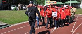 Dzień. Na bieżni boiska sportowego maszerują kolejności. Druh OSP ubrany w mundur galowy ze sznurem, rogatywkę oraz buty z opinaczami. Za nim podąża grupa dzieci ubranych w identyczne spodnie w kolorze granatowym, czerwone bluzy oraz czerwone czapki z daszkiem. Za nimi podąża grupa kolejnych dzieci. W tle dwa namioty pokryte zielonym materiałem. Z lewej strony grupa osób przygląda się przemarszowi dzieci