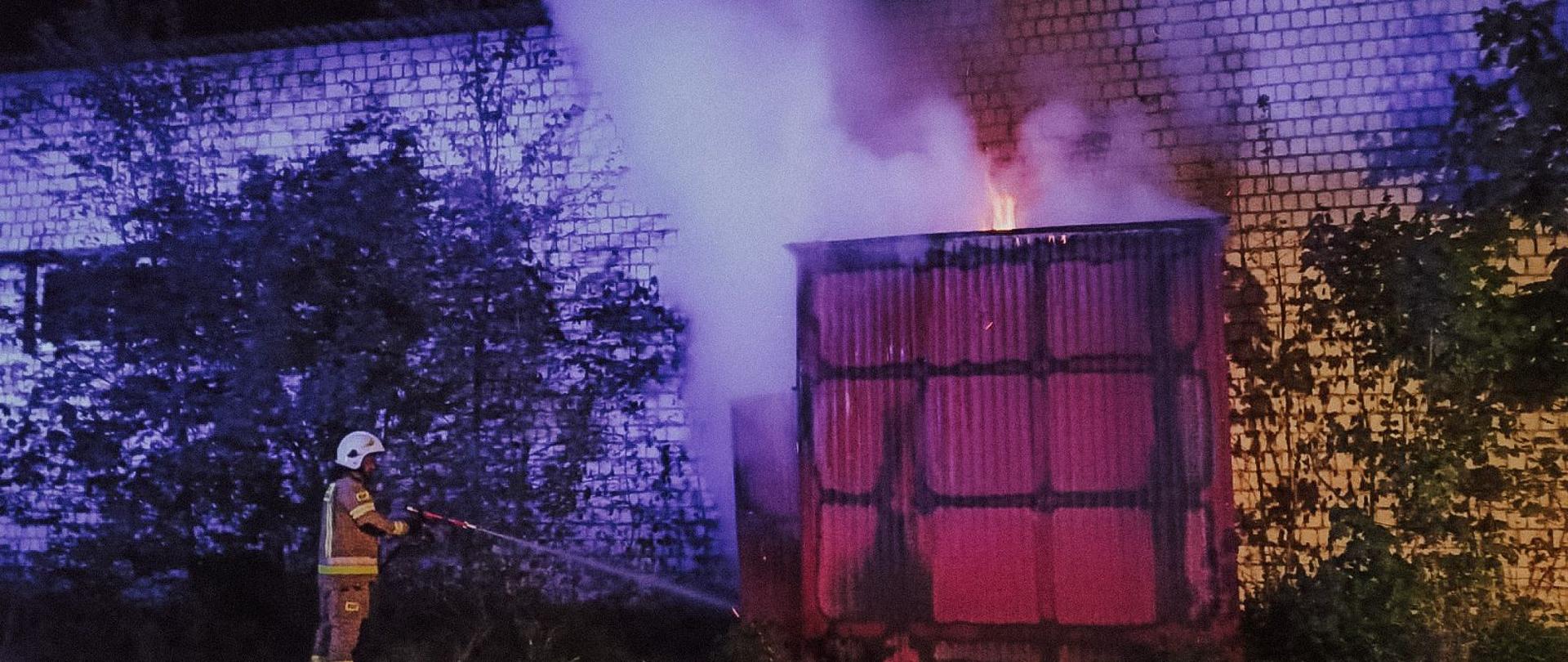 Strażak gasi palący się obiekt. W pobliżu krzewy i zabudowania. Zdjęcie wykonane w porze nocnej.