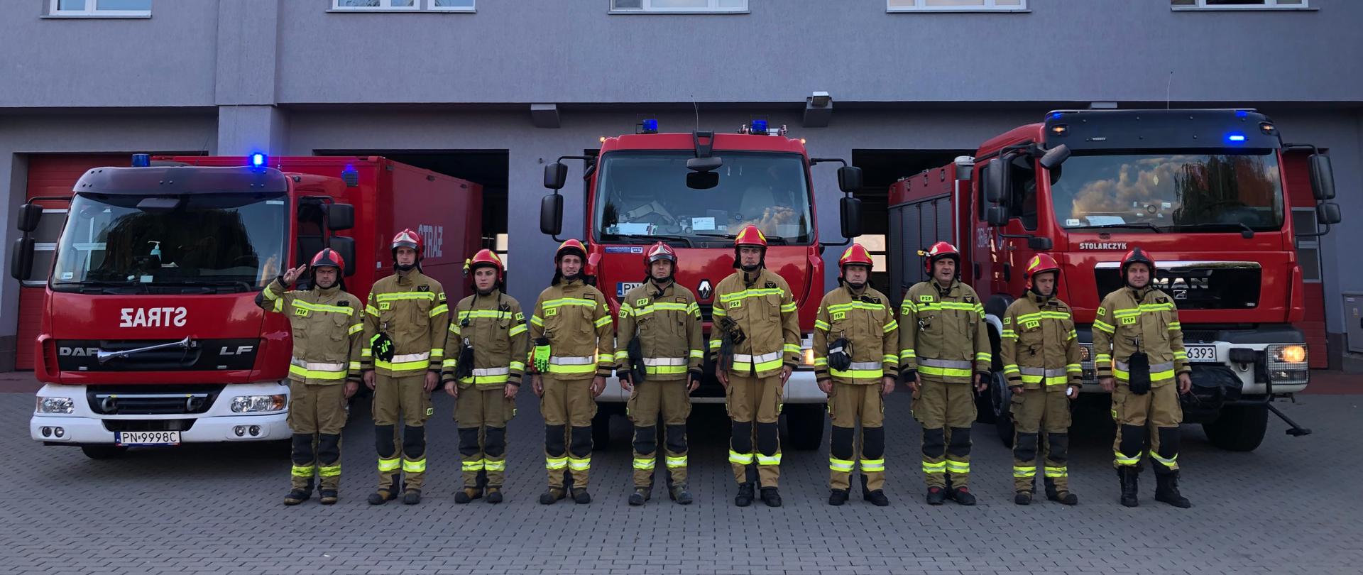Zdjęcie przedstawia strażaków stojących przed garażem podczas oddawania hołdu zmarłemu strażakowi. Strażacy ubrani w ubrania specjalne. Za nimi stoją samochody strażackie z włączonymi sygnałami świetlnymi. 