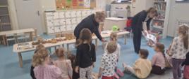 Dzieci siedzą na podłodze w sali przedszkolnej i uczestniczą w zajęciach o ziemniakach. Dwie prowadzące prezentują materiały edukacyjne, a na stolikach leżą ziemniaki różnych odmian, koszyki i ilustracje związane z ich uprawą.