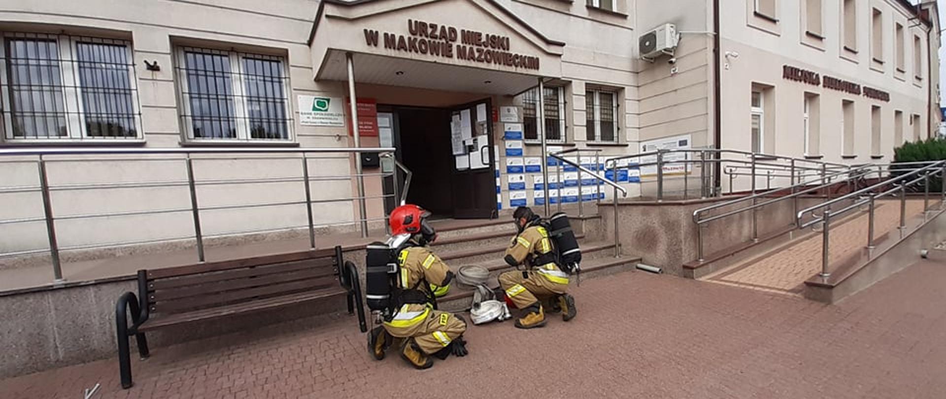 Strażacy zakładają aparaty ochrony dróg oddechowych przed wejściem do budynku Urzędu Miasta.