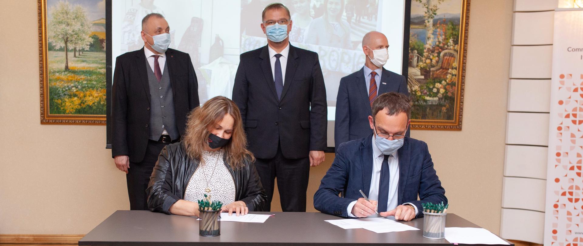 trwa ceremonia podpisania umowy - kobieta i mężczyzna podpisują dokumenty, trzech mężczyzn stoi za podpisujacymi dokumentami