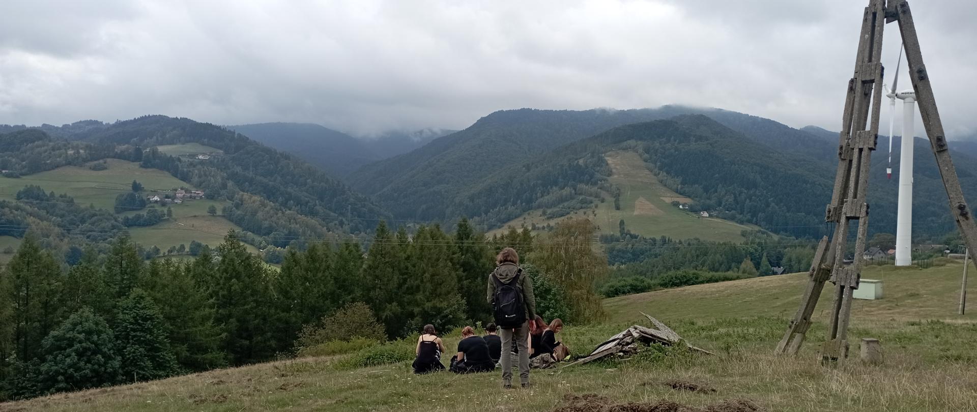 Uczniowie wraz z nauczycielem uczestniczą w zajęciach plenerowych na górskiej łące.
W tle rozciąga się panorama zalesionych beskidzkich wzgórz spowitych chmurami.
Po prawej stronie zdjęcia widoczna jest turbina wiatrowa.