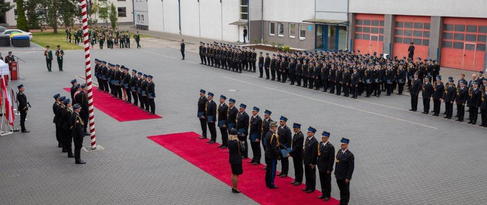 Zdjęcie wykonane w ciągu dnia, przedstawia pododdziały aspirantów Państwowej Straży Pożarnej podczas promocji na placu Szkoły Aspirantów Państwowej Straży Pożarnej w Poznaniu. W tle orkiestra oraz budynki, garaże oraz hala sportowa Szkoły Aspirantów. 