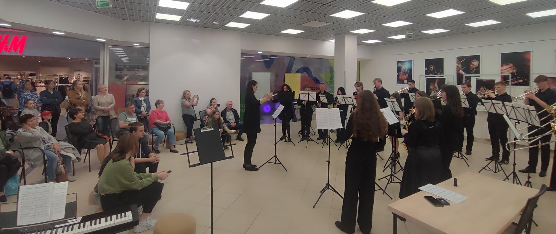 W jasnej dużej sali po prawej stronie stojąc przy pulpitach gra orkiestra na różnych instrumentach dętych, w środku stoi kobieta i dyryguje, po lewej stronie stoi i siedzi na krzesłach publiczność.