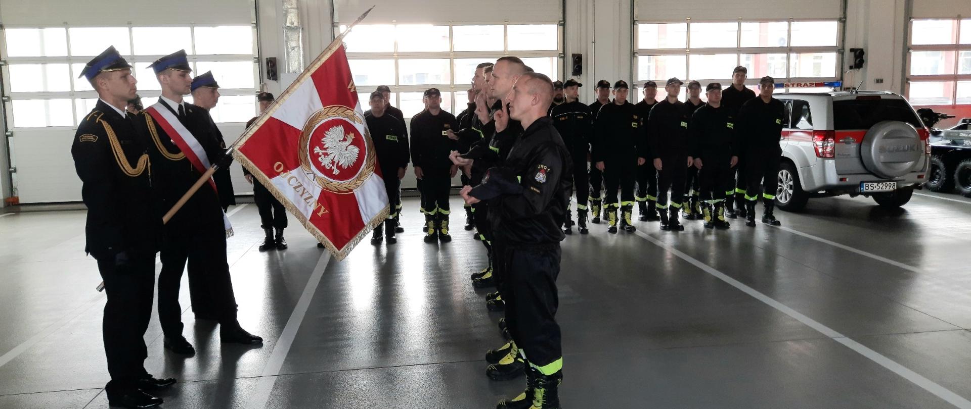 Zdjęcie przestawia strażaków w garażu jeden z nich trzyma sztandar a kilku ma podniesione prawe dłonie do przysięgi