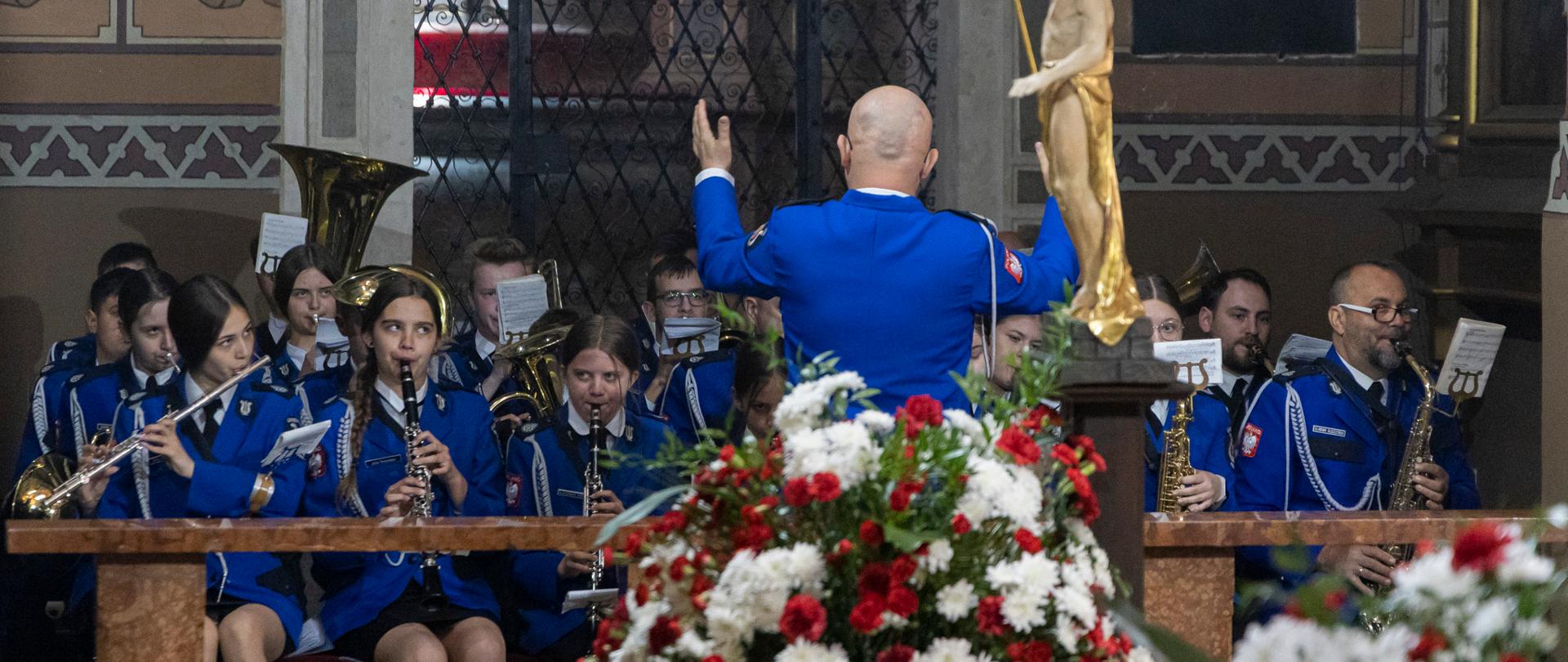 Tyłem do obiektywu a przodem do orkiestry stoi kapelmistrz. Ręce trzyma w górze, dyryguje. Dalej widać orkiestrantów z instrumentami. Na pierwszym planie postaci przysłaniają ozdobne kandelabry z kwiatów.