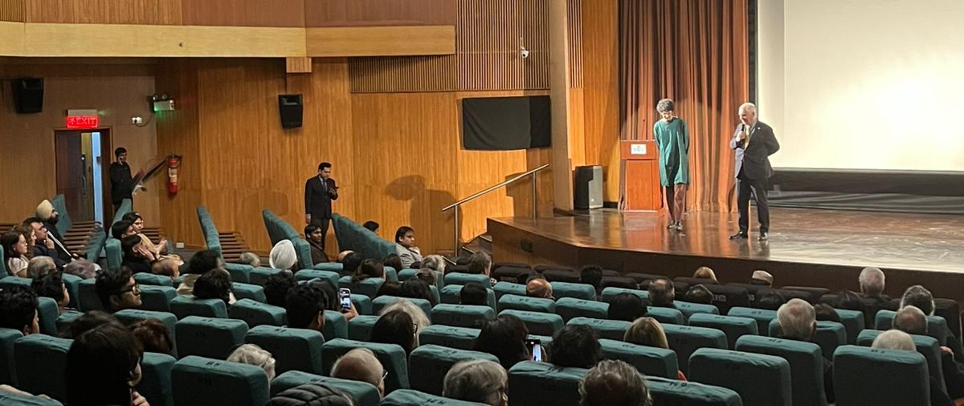 A cinema hall full of people during the screening of the film Letters from Wolf Street. On stage, the Head of Mission Piotr Świtalski and the film's director Arjun Talwar are speaking.