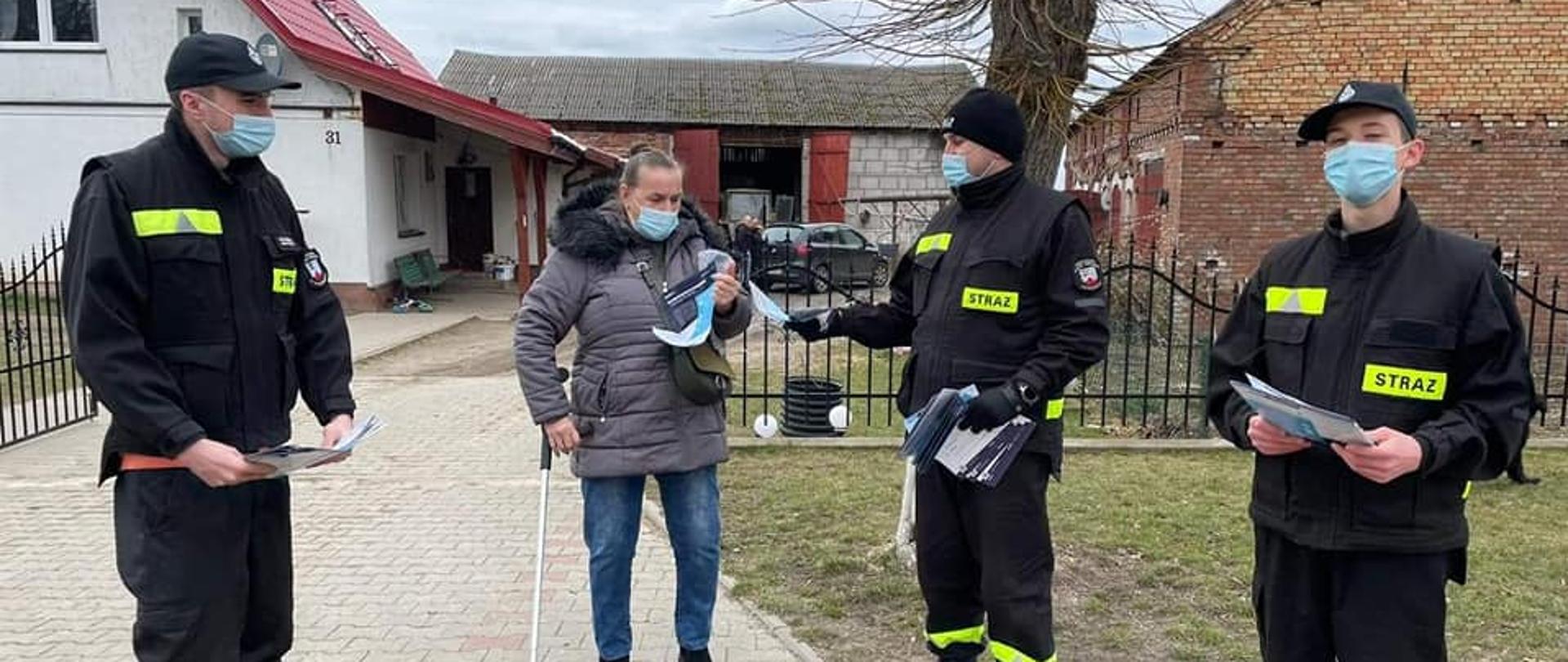 Na zdjęciu znajdują się strażacy, którzy roznoszą ulotki. Ubrani są w czarne ubranie koszarowe oraz maseczki ochronne. Stoją przy ulicy wręczając mieszkance miejscowości ulotkę zachęcającą do szczepień przeciw COVID-19. W tle znajduje się drzewo oraz podwórko gospodarstwa. Zdjęcie wykonano w dzień.