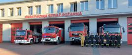 Strażacy przed samochodami na tle Komendy Powiatowej
