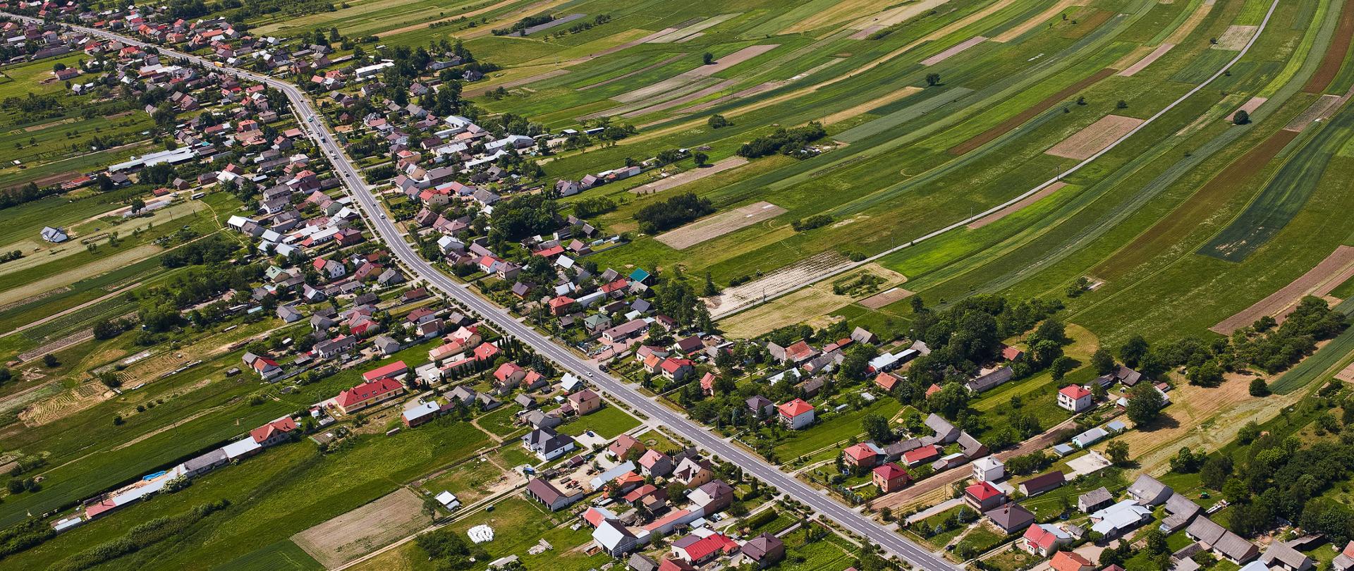 Widok z lotu ptaka na wiejską miejscowość położoną wśród rozległych pól uprawnych. Przez środek zdjęcia biegnie główna droga, wzdłuż której stoją domy jednorodzinne i zabudowania gospodarcze. Otaczające wieś pola tworzą mozaikę długich, wąskich pasów w różnych odcieniach zieleni i brązu. Całość przedstawia spokojny, rolniczy krajobraz.