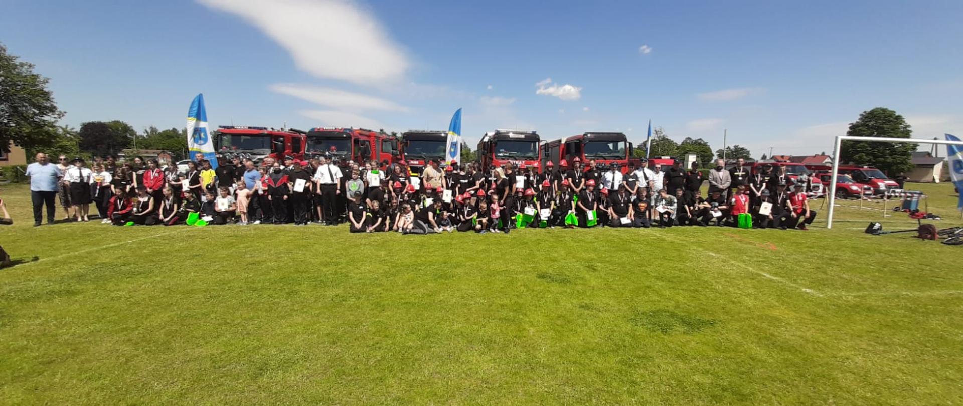 Na stadionie sportowym w Czarnem stoją wszystkie drużyny dziecięce wraz z opiekunami i organizatorami na tle samochodów pożarniczych.