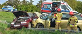 Fotografia przedstawia pojazdy po kolizji. Widać także strażaków PSP wykonujących jakieś czynności. W tle widać przybyły na miejsce zdarzenia pojazd ZRM.