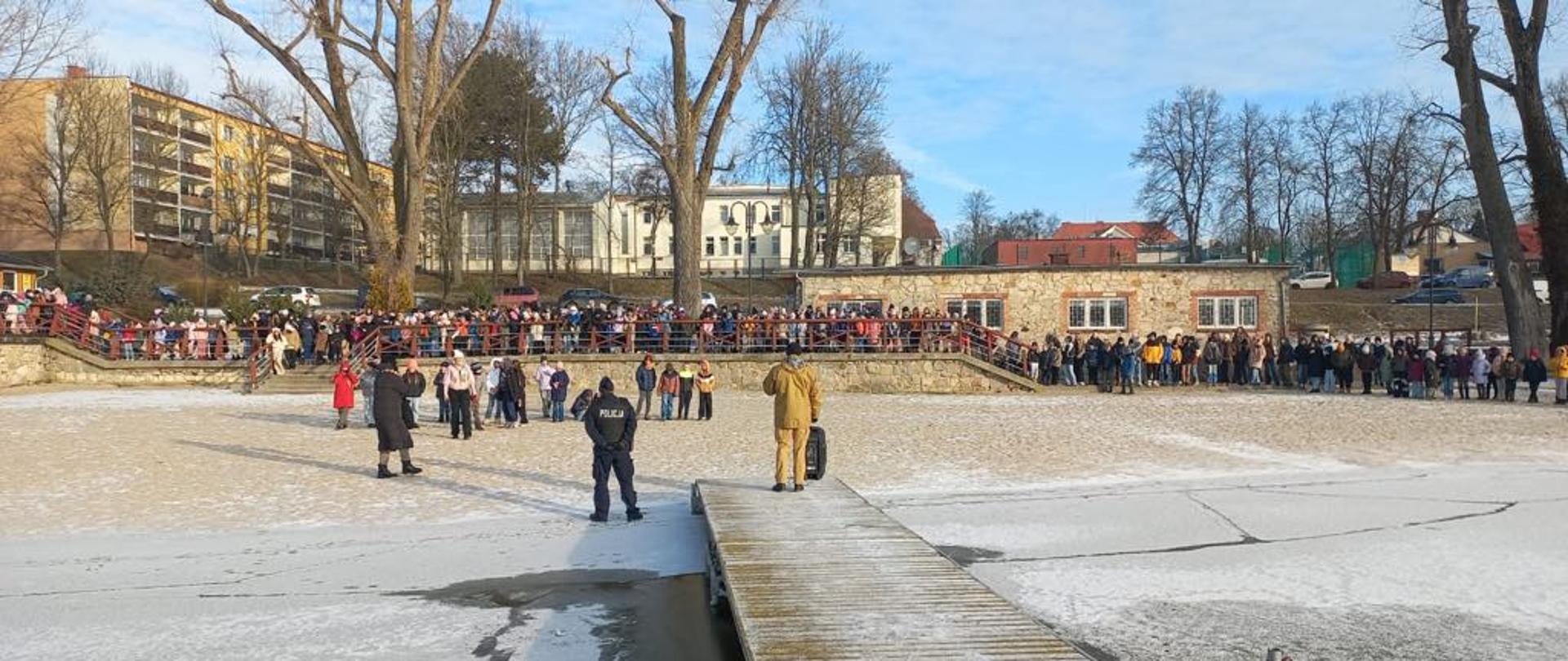Plaża miejska. w środku kadru stoi Policjant i strażak. Przed nimi na promenadzie stoją dzieci z nauczycielami
