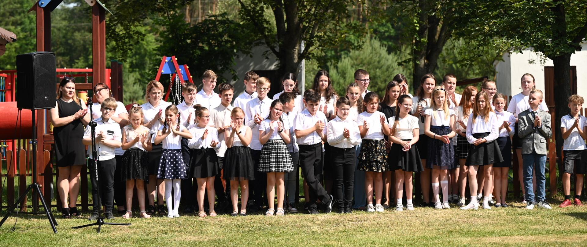Elegancko ubrane dzieci stoją rzędem na dworze, obok plac zabaw.