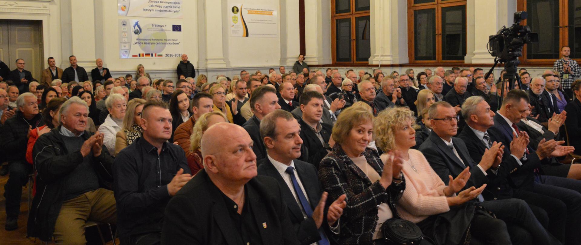 Mieszkańcy powiatu prudnickiego siedzą i klaszczą w dłonie podczas przemówienia ministra