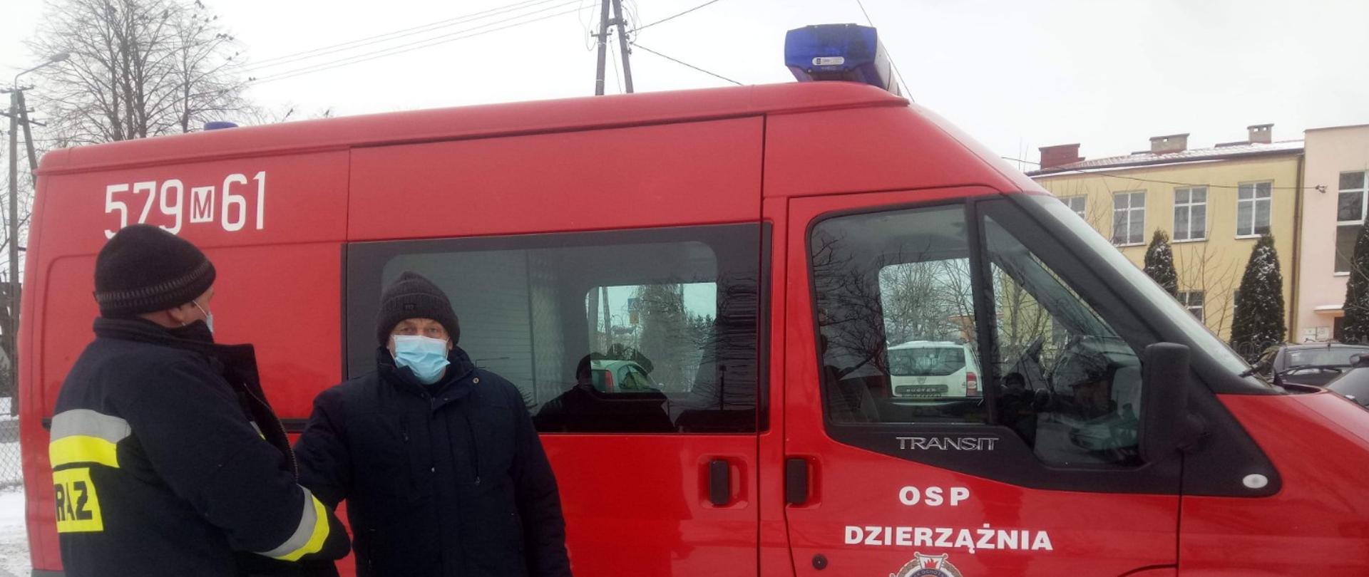 Strażak ubrany w ciemny mundur ze starszym Panem ubranym w czarną kurtkę i czarne spodnie. W tle znajduje się czerwony samochód strażacki.