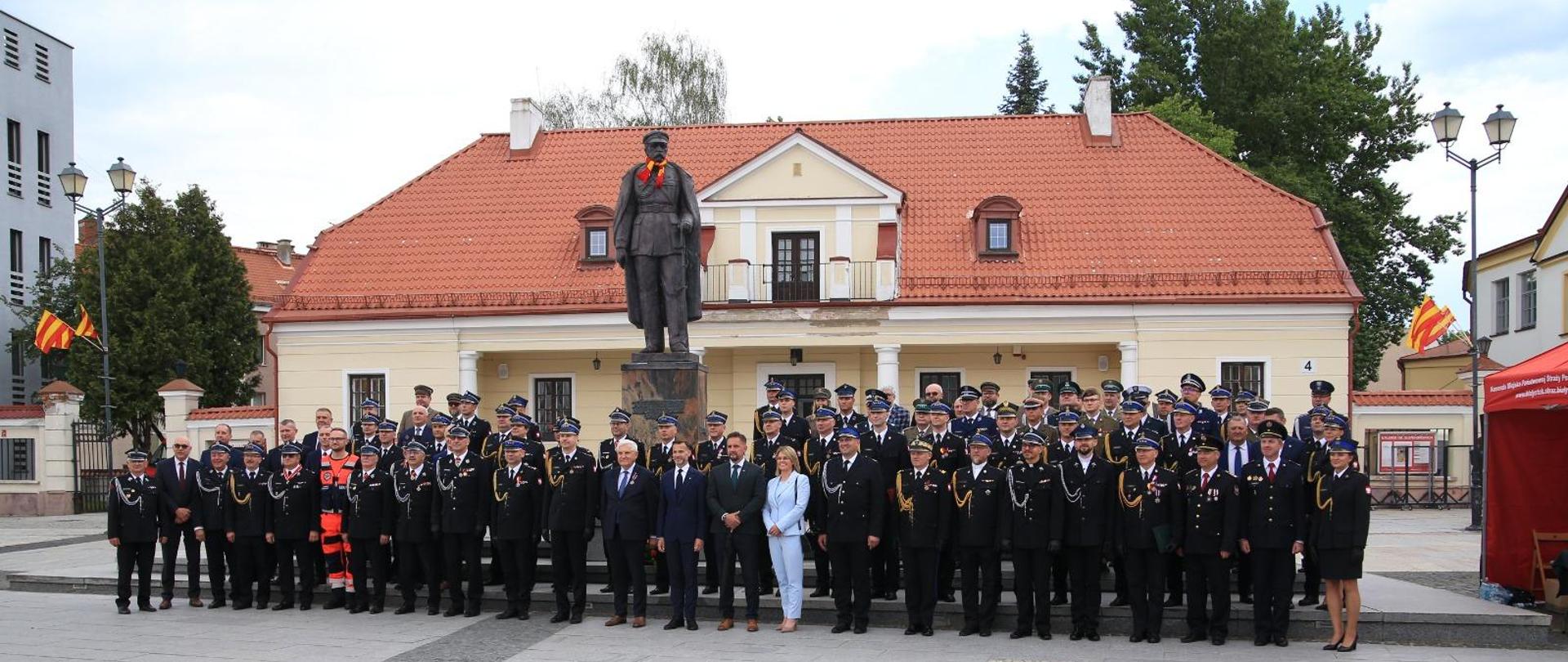 Pamiątkowa fotografia uczestników uroczystości strażackich. Zaproszeni goście stoją na placu przy pomniku Marszałka Józefa Piłsudskiego w Białymstoku. Za osobami stojącymi na zdjęciu widoczny pomnik Józefa Piłsudskiego.