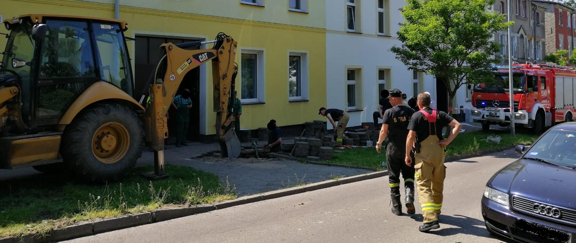 Zdjęcie przedstawia chodnik a w nim kopią dziurę robotnicy ubrani na zielono. Również kopie żółta koparka. 