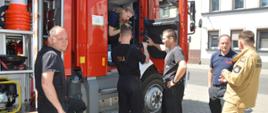 Wizyta strażaków z Attendorn. Plac manewrowy przed komendą. Komendant powiatowy oraz strażacy ze zmiany służbowej prezentują gościom wóz ratowniczo-gaśniczy i sprzęt. W tle udynek mieszkalny.