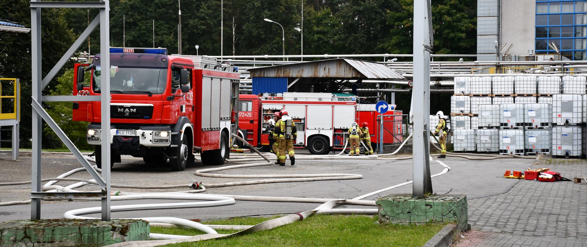 Na zdjęciu widzimy samochody pożarnicze oraz strażaków w pełnym uzbrojeniu, którzy rozwijają linie gaśnicze. W tle widać budynki zakładów oraz zbiorniku z chemikaliami.