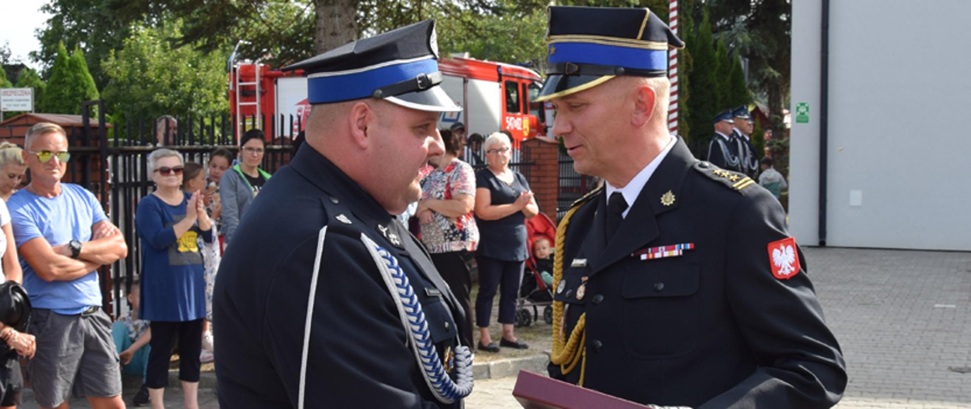 Dwóch strażaków stoi na przeciwko siebie. Podają sobie ręce. Jeden z nich trzyma w dłoni bordowe pudełko. Obaj są lekko uśmiechnięci. W tle widać kobiety i mężczyznę którzy się przyglądają. Drzewa i samochód strażacki 